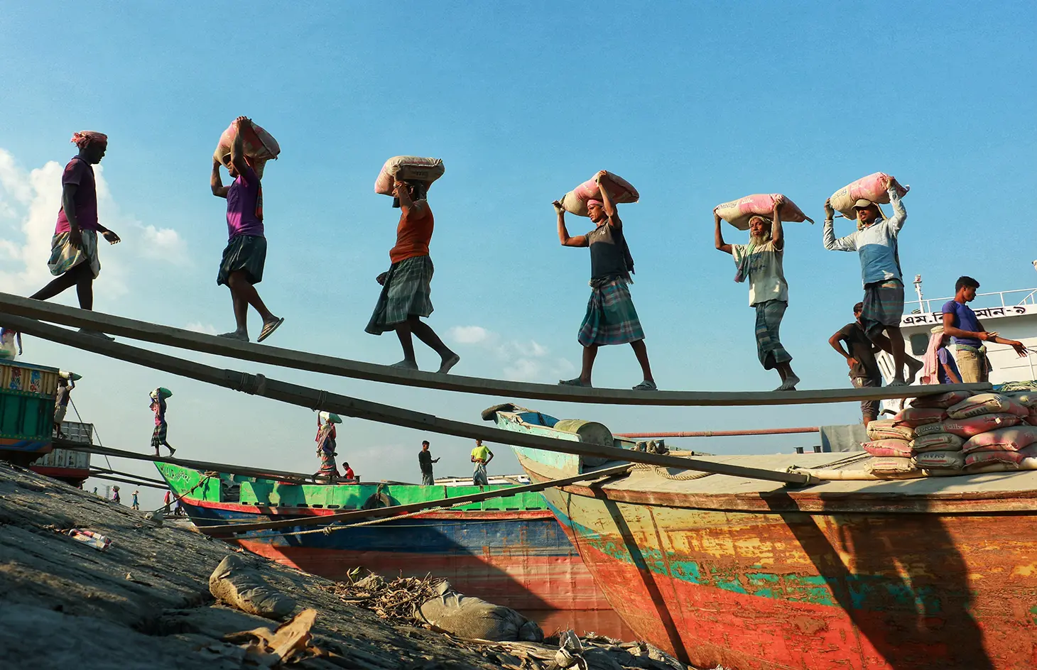 A photo of a group of Asian workers walking along a plant from a boat to the shore, carrying sacks on their heads.