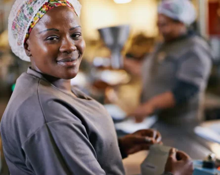 A photo of a smiling black woman wearing a hair net, working in a food factory.